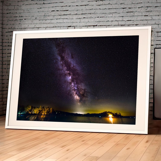 A vivid Milky Way core glows in a vertical sweep across a dark, starry night sky. The stars and galactic dust create a textured ribbon of light. Below, a calm lake mirrors faint reflections of the stars, with a rocky shoreline in the foreground and a glowing island of trees lit in the distance.