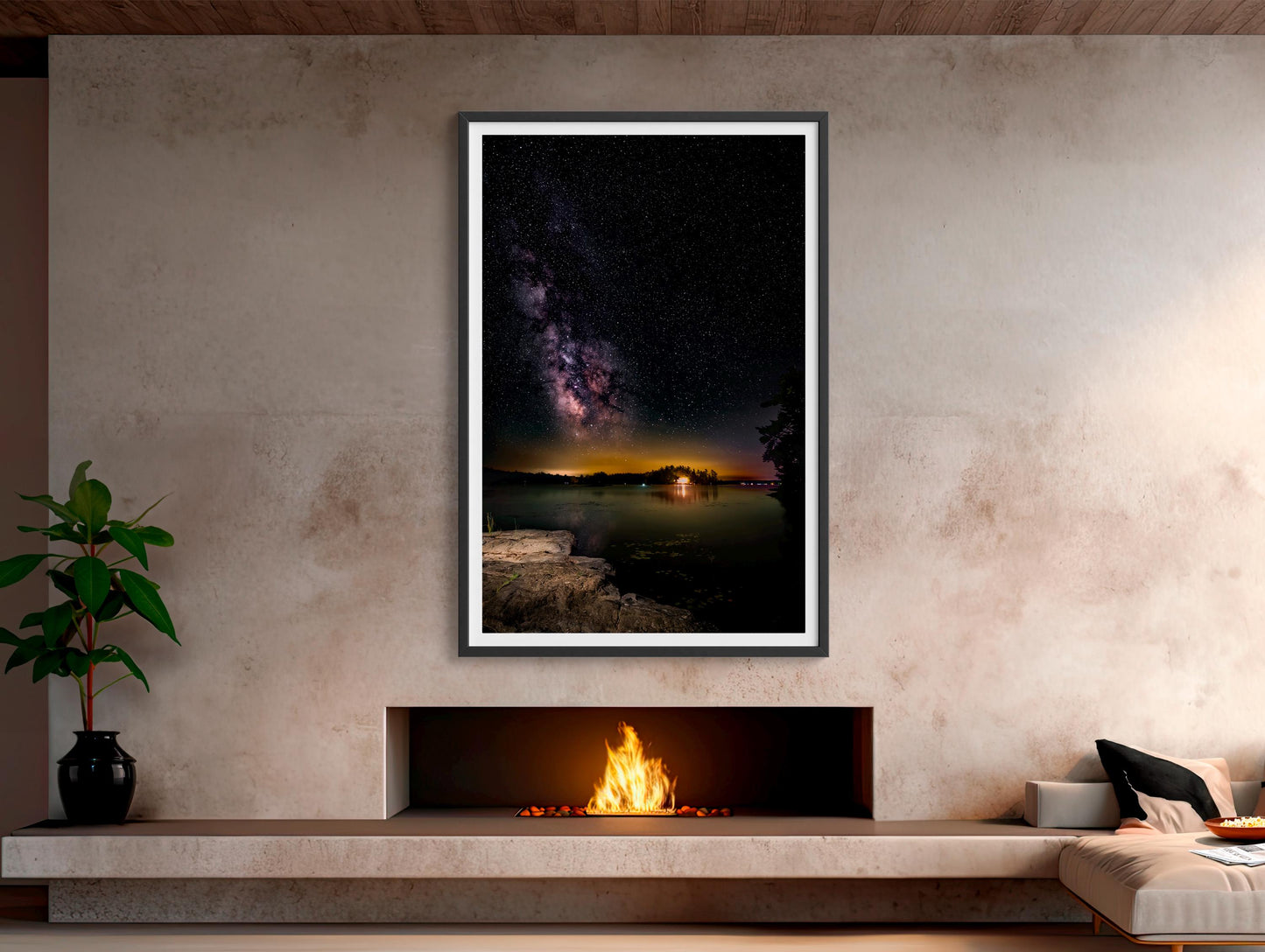 A vertical photograph of the Milky Way core arcs high above a calm, dark lake. A flat rock extends into the water, serving as the photographer’s platform. Countless stars and cosmic dust form a glowing band of light, reflecting faintly on the water’s surface under the deep black sky.