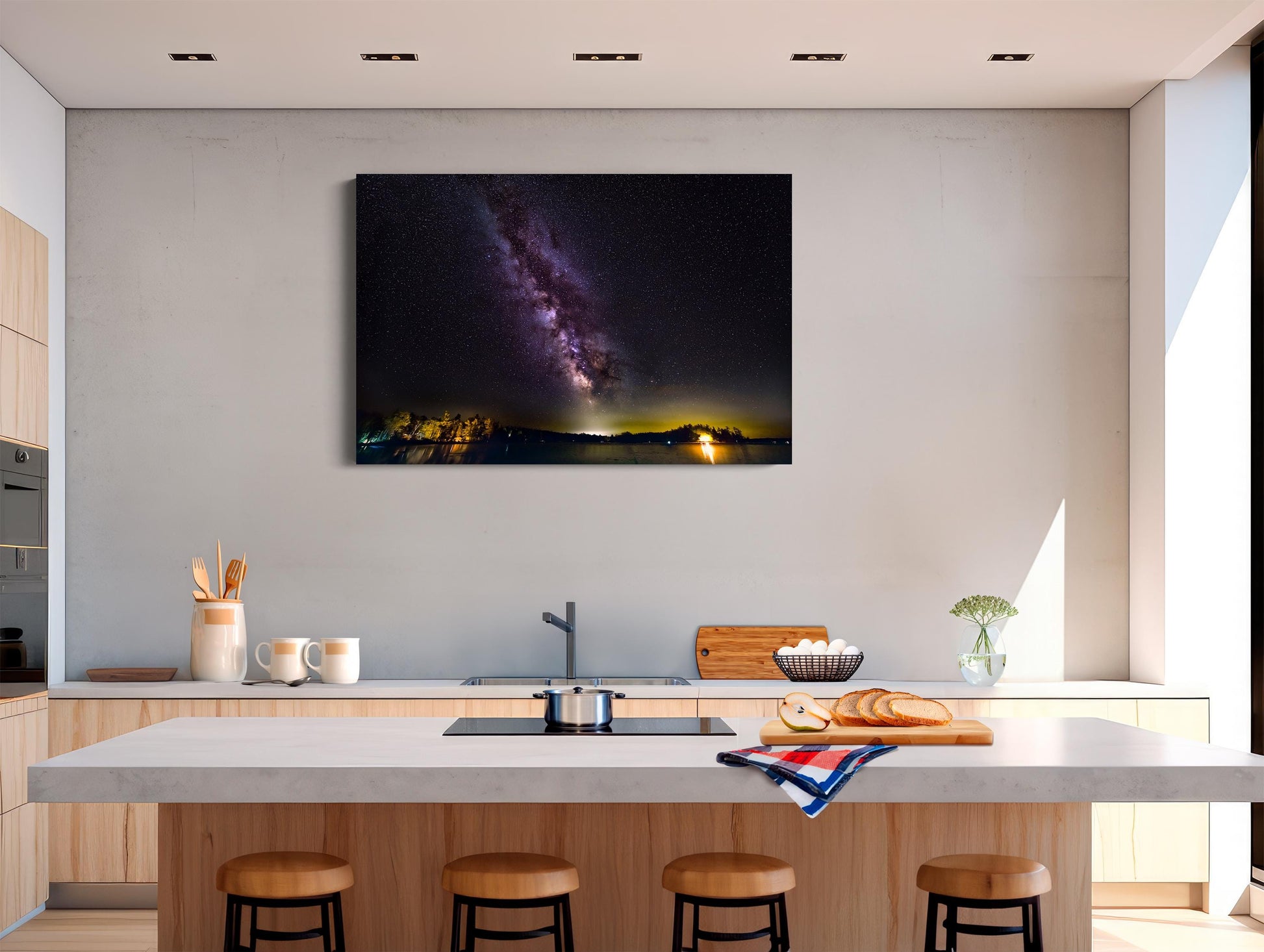 A vivid Milky Way core glows in a vertical sweep across a dark, starry night sky. The stars and galactic dust create a textured ribbon of light. Below, a calm lake mirrors faint reflections of the stars, with a rocky shoreline in the foreground and a glowing island of trees lit in the distance.