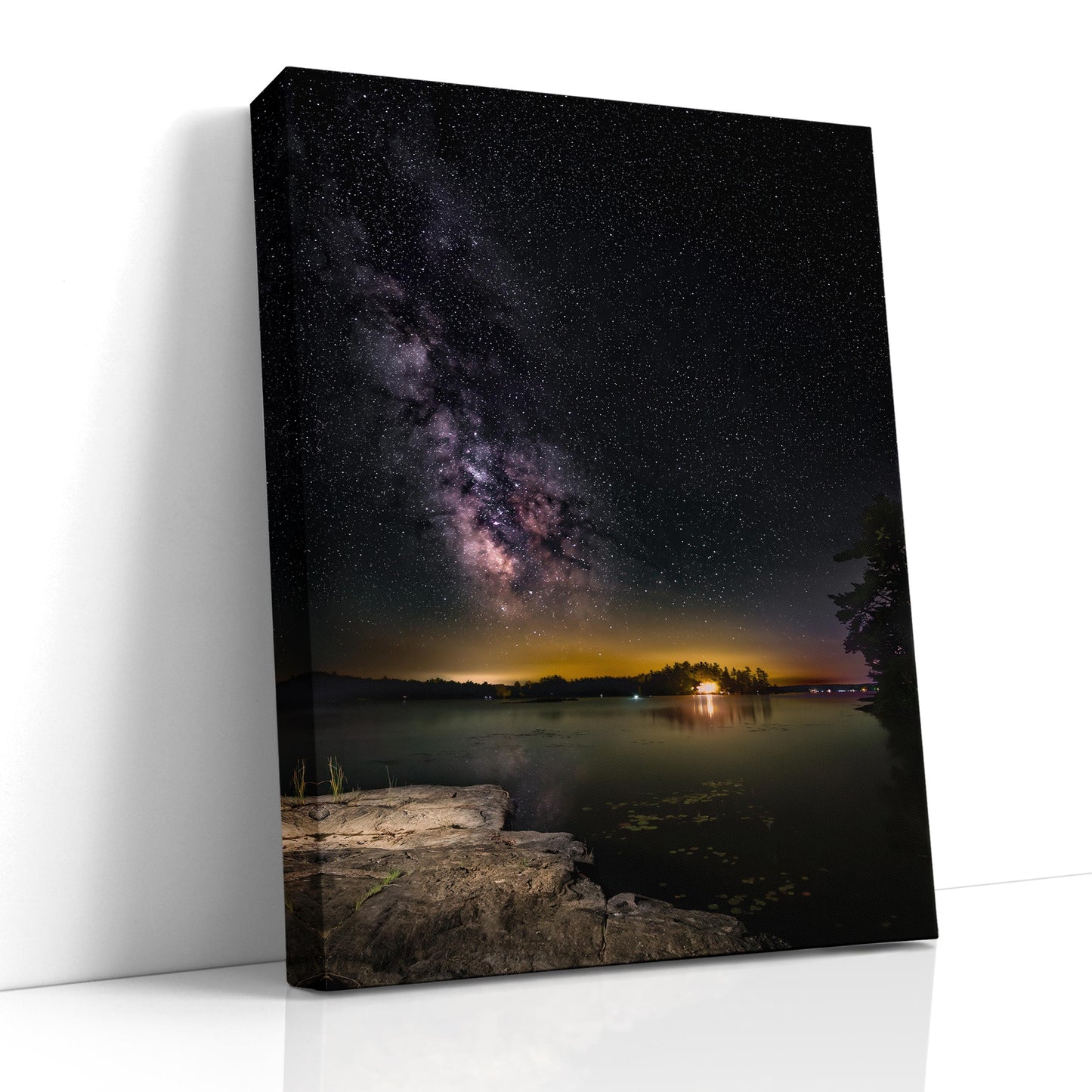 A vertical photograph of the Milky Way core arcs high above a calm, dark lake. A flat rock extends into the water, serving as the photographer’s platform. Countless stars and cosmic dust form a glowing band of light, reflecting faintly on the water’s surface under the deep black sky.