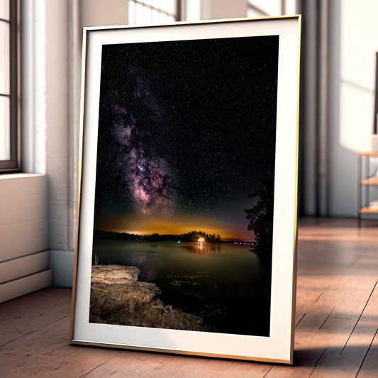 A vertical photograph of the Milky Way core arcs high above a calm, dark lake. A flat rock extends into the water, serving as the photographer’s platform. Countless stars and cosmic dust form a glowing band of light, reflecting faintly on the water’s surface under the deep black sky.
