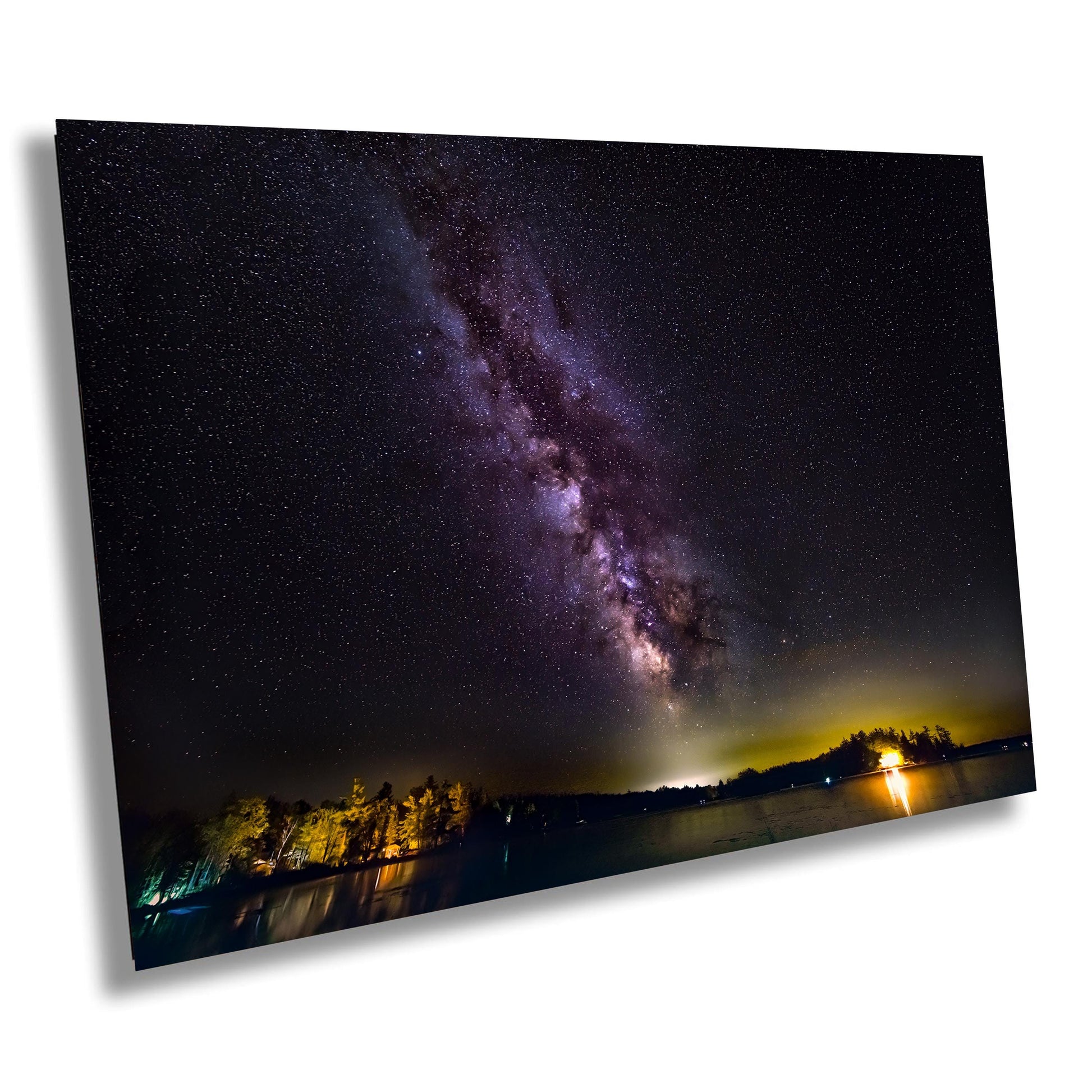 A vivid Milky Way core glows in a vertical sweep across a dark, starry night sky. The stars and galactic dust create a textured ribbon of light. Below, a calm lake mirrors faint reflections of the stars, with a rocky shoreline in the foreground and a glowing island of trees lit in the distance.